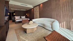 A modern hotel room with a curved sofa, a round coffee table, a bed with white linens, and a large wall mural. - Mavilu Luxury Hotel (Bodrum)