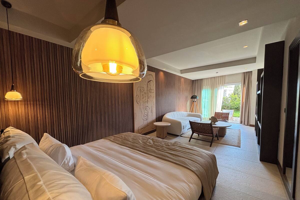 A modern bedroom with a large bed, a desk, and a chair. There is a window with curtains and a view of greenery outside.