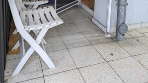 A small porch with a white chair and a table, a window with curtains, and a metal pipe on the wall.