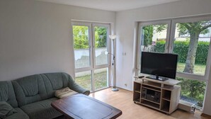 A living room with a green sofa, a wooden coffee table, a TV on a wooden stand, and large windows with a view of greenery.