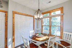 A dining area with a round table, white chairs, a chandelier, and large windows with a view of trees.