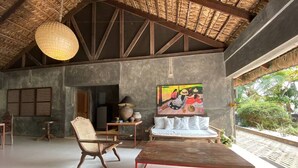 Une pièce spacieuse avec un plafond haut, une grande table en bois, un canapé et un fauteuil traditionnel en osier. Un tableau orne le mur, et on aperçoit un toit de chaume.
