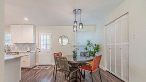 A compact kitchen with white cabinets, a dining table with four chairs, a hanging light fixture, and a window with blinds.