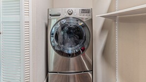 A stainless steel front-loading washing machine in a laundry room with a white wall and a white shelf above it.