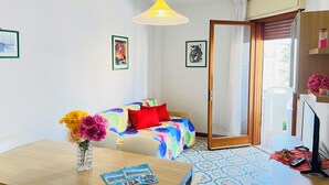 Een kamer met een bed, een tafel met een bloevas, een bureau met een computer, en een deur die naar buiten leidt.