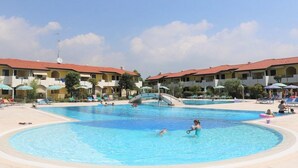 A swimming pool area with multiple pools, lounge chairs, umbrellas, and a bridge connecting the pools. - Lovely flat with garden (Bibione)