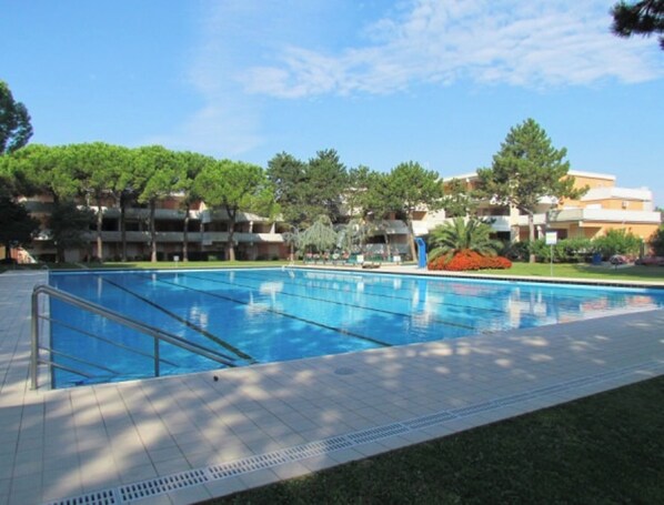 A swimming pool with a tiled deck, surrounded by greenery and trees, with a multi-story building in the background.