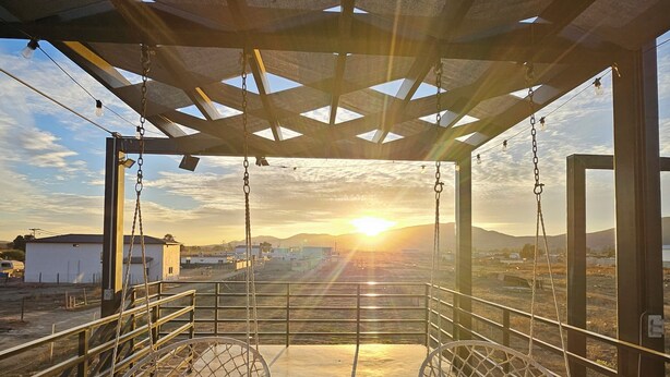 Un espace extérieur aménagé, abrité, avec deux fauteuils suspendus, un sol en béton et une vue sur le coucher de soleil se dégageant sur un paysage urbain.