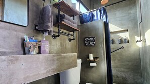 Une salle de bain avec un mur en béton, des toilettes blanches, une étagère avec des articles de toilette et un panneau indiquant qu'il ne faut pas jeter le papier toilette dans les toilettes.