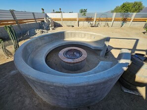 A circular concrete structure with a central fire pit, surrounded by a sandy area with construction materials and a metal fence in the background. - 
“A Cabin Made for Escapes, Not Just Stays.” (Ejido El Porvenir (Guadalupe))