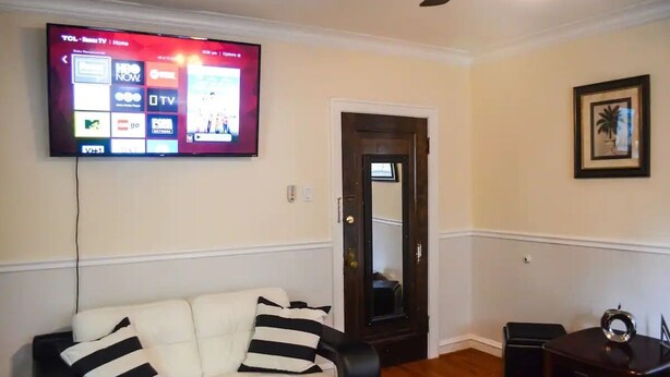 A living room with a white couch, a flat-screen TV mounted on the wall, a wooden door, and a framed picture on the wall.