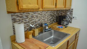 A kitchen with wooden cabinets, a tiled backsplash, a stainless steel sink, a cutting board, a paper towel holder, a knife block, and a basket of items on the countertop.