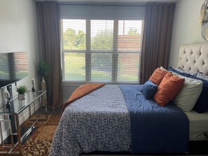 Ein Schlafzimmer mit Bett, Fernseher, Pflanze und einem Fenster mit Jalousien.