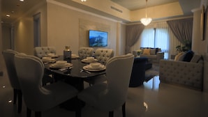 A dining area with a round table set for six, surrounded by upholstered chairs. There is a chandelier above the table and a television mounted on the wall.