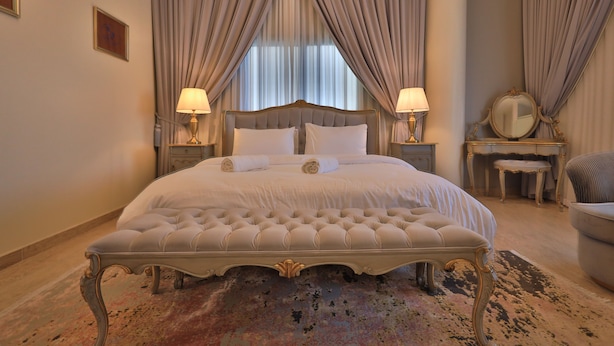 A large bed with a tufted headboard, two bedside lamps, a small table with a mirror, and a chair.