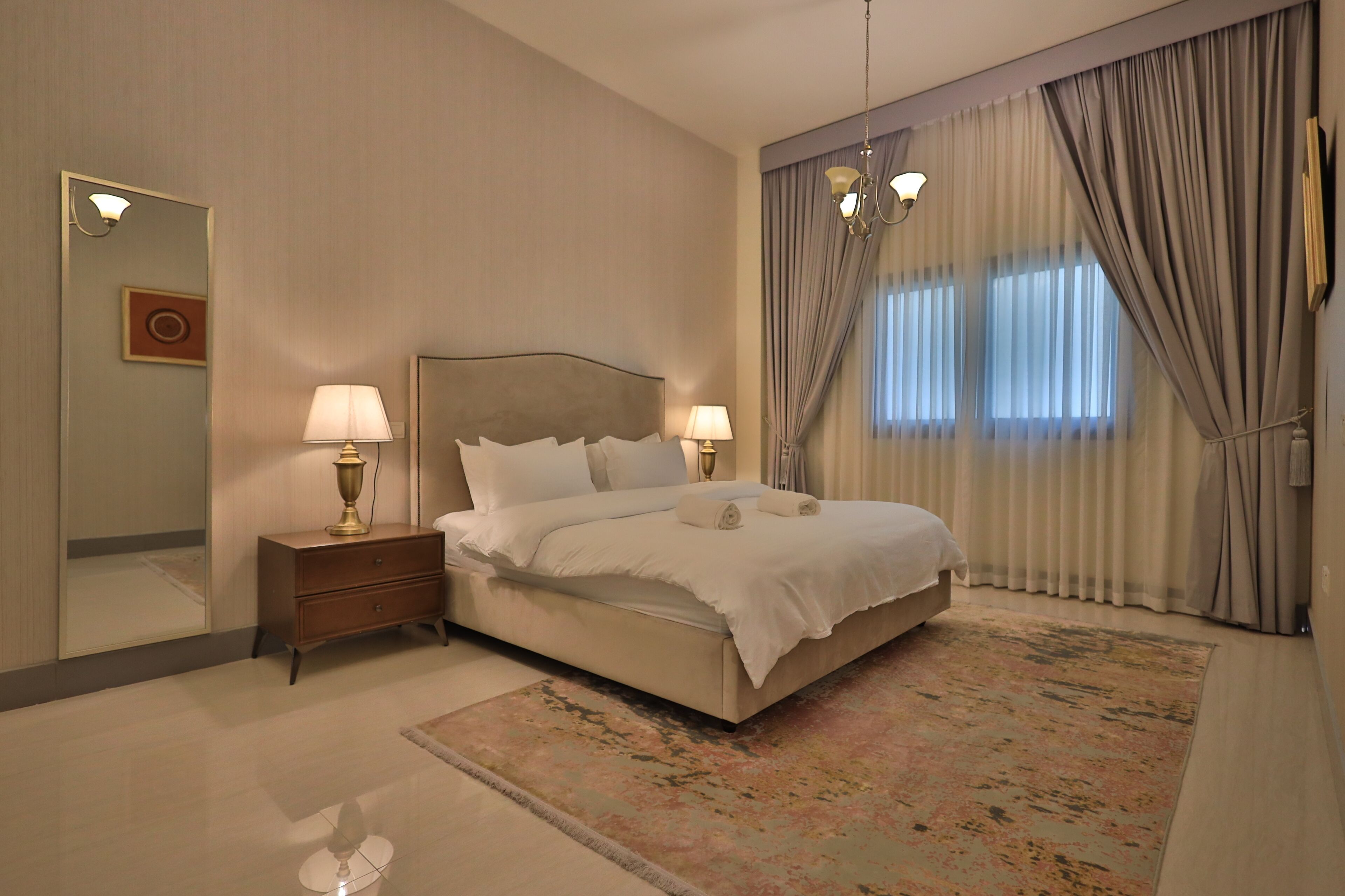 A bedroom with a large bed, two bedside tables with lamps, a mirror, and a window with curtains.