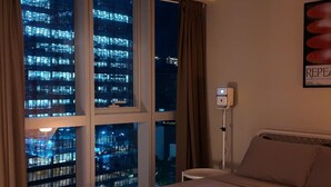A hotel room with a large bed, a night view of a cityscape through the windows, a bedside table with a lamp, and a patterned rug.