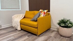 A mustard-colored sofa with a beige throw blanket and colorful pillows, positioned next to a potted plant with white flowers.