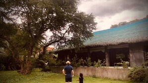 Un hombre y un niño de pie en un área con césped, con un edificio de techo de paja al fondo.