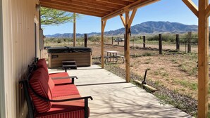 Une terrasse couverte avec un plafond en bois, un canapé à rayures rouges, une table et un jacuzzi. La terrasse offre une vue sur un paysage désertique avec des montagnes à l’horizon.