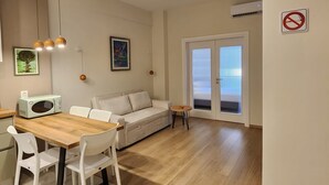 Uma sala de estar moderna com uma mesa de jantar de madeira, cadeiras brancas, um sofá bege e uma pequena mesa lateral. Há um quadro na parede, uma luminária no teto e um ar condicionado.