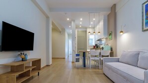 Uma sala moderna com televisão de tela plana, prateleiras de madeira, um sofá, uma área de refeições com cadeiras e uma cozinha ao fundo.