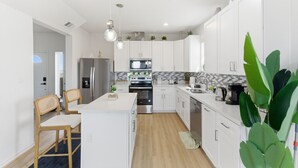 A modern kitchen with white cabinets, a central island, wooden chairs, and a large green plant.