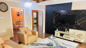 Una sala de estar con sofá, sillón y una mesa de centro sobre un tapete con diseño de mármol. Un televisor de pantalla plana está montado en una pared negra, y se ve un comedor al fondo.
