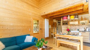 A wooden cabin interior with a blue sofa, wooden dining table, and a kitchen area with cabinets and a refrigerator. - The Bear Fountain (LA FECLAZ LES DESERTS)