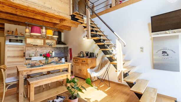 A modern kitchen with wooden cabinetry, a dining area with a wooden table and chairs, and a staircase with a metal railing. - The Bear Fountain (LA FECLAZ LES DESERTS)