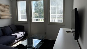 A modern living room with a glass coffee table, a dark sofa, and a flat-screen TV mounted on the wall. There are three windows with blinds, and a wooden slat shelf is visible on the left wall.