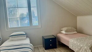 A bedroom with a sloped ceiling, a window with blinds, a bed with striped bedding, a bedside table with a drawer, and a bedside lamp.