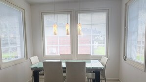 A dining area with a glass table and four chairs, large windows with blinds, and a light fixture above.