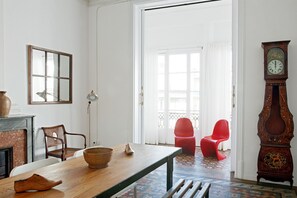 Een kamer met een houten tafel, stoelen, een klok en uitzicht door een geopende deur.