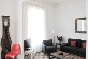 A living room with a large window, a clock, a television, a sofa, a chair, and a coffee table. - Modernist apartment with AC in central Barcelona! (Barcelona)