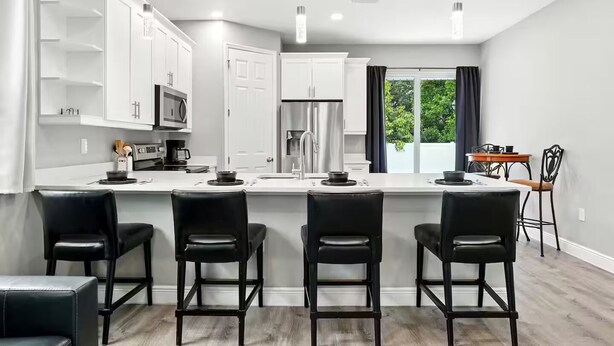 A modern kitchen with white cabinets, a central island with bar stools, and a dining area with a table and chairs. - Entire New Home w/1 King, 3 Queen beds and Fenced backyard (Bonita Springs)