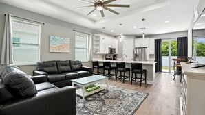 A modern living room with a black leather sofa, a coffee table, and a patterned rug. There is a kitchen area with white cabinets and a stainless steel refrigerator. - Entire New Home w/1 King, 3 Queen beds and Fenced backyard (Bonita Springs)