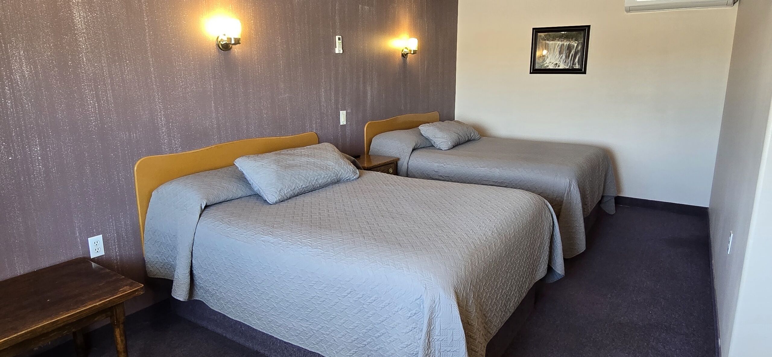 A hotel room with two beds, each with a headboard and pillows, a bedside table, and wall-mounted lights.