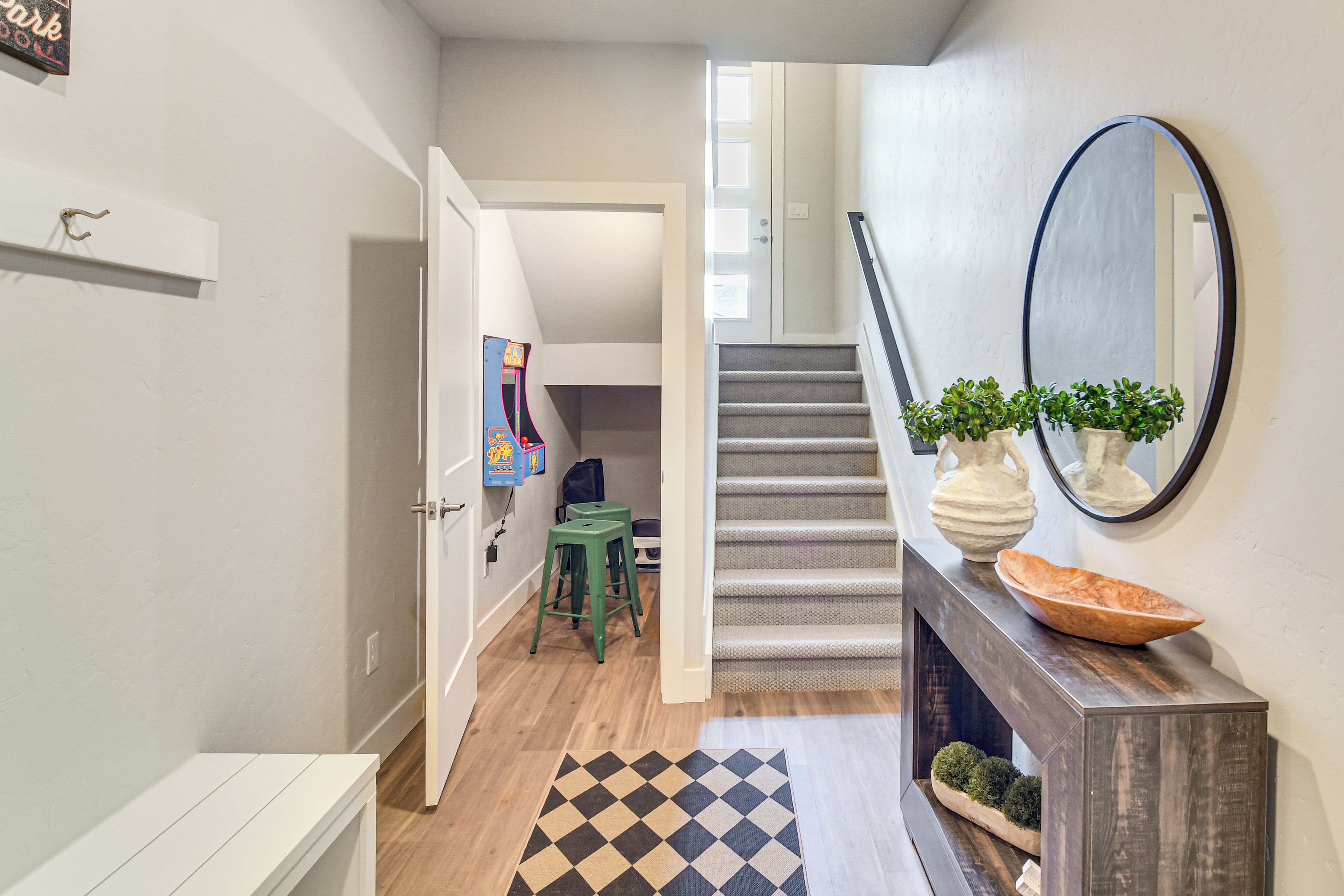 Un couloir avec un escalier, un plancher en bois, un tapis à carreaux, un tabouret vert et un miroir posé sur une console.