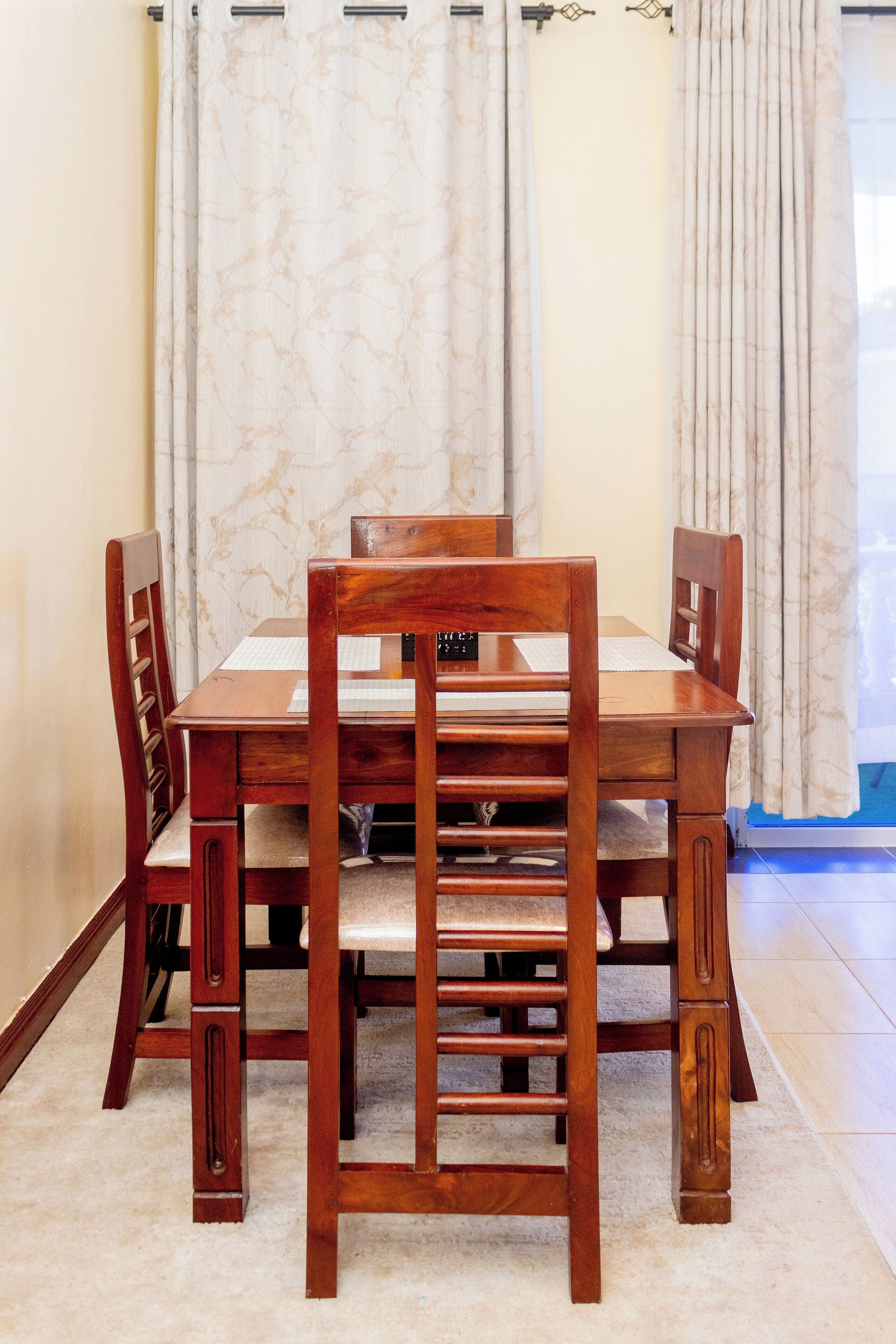 Una mesa de comedor de madera con cuatro sillas, un mantel blanco y una cortina con estampado de mármol.