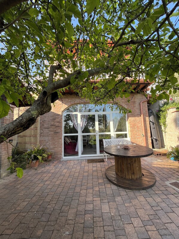 A brick patio with a wooden table and chairs, surrounded by potted plants and a large window.