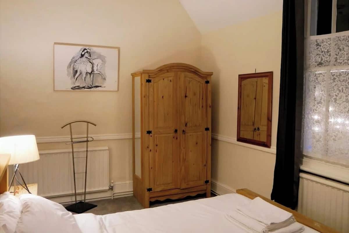 A bedroom with a bed, a wooden wardrobe, a radiator, a floor lamp, and a window with lace curtains.