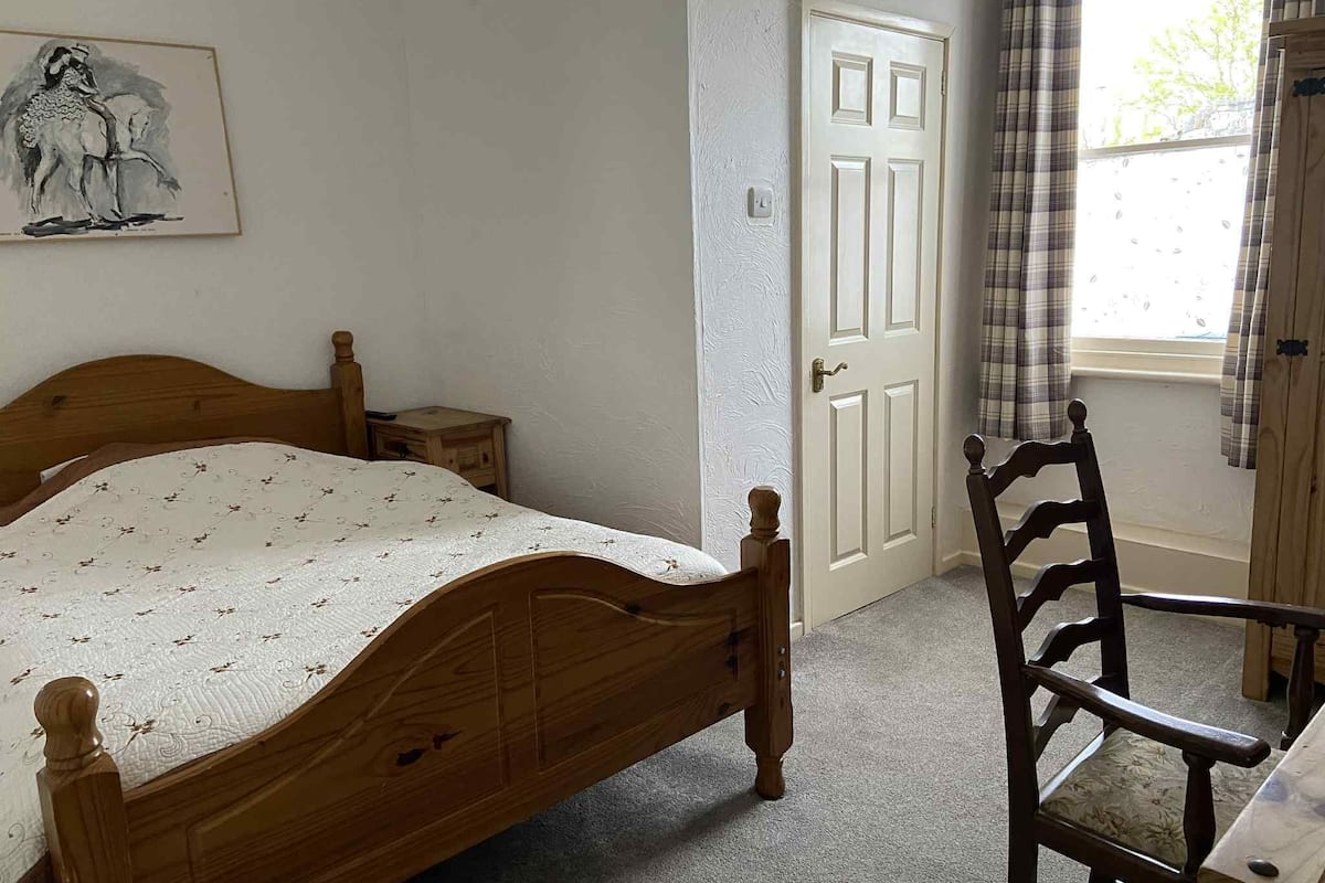 A bedroom with a wooden bed, a small bedside table, a chair, a door, and a window with curtains.