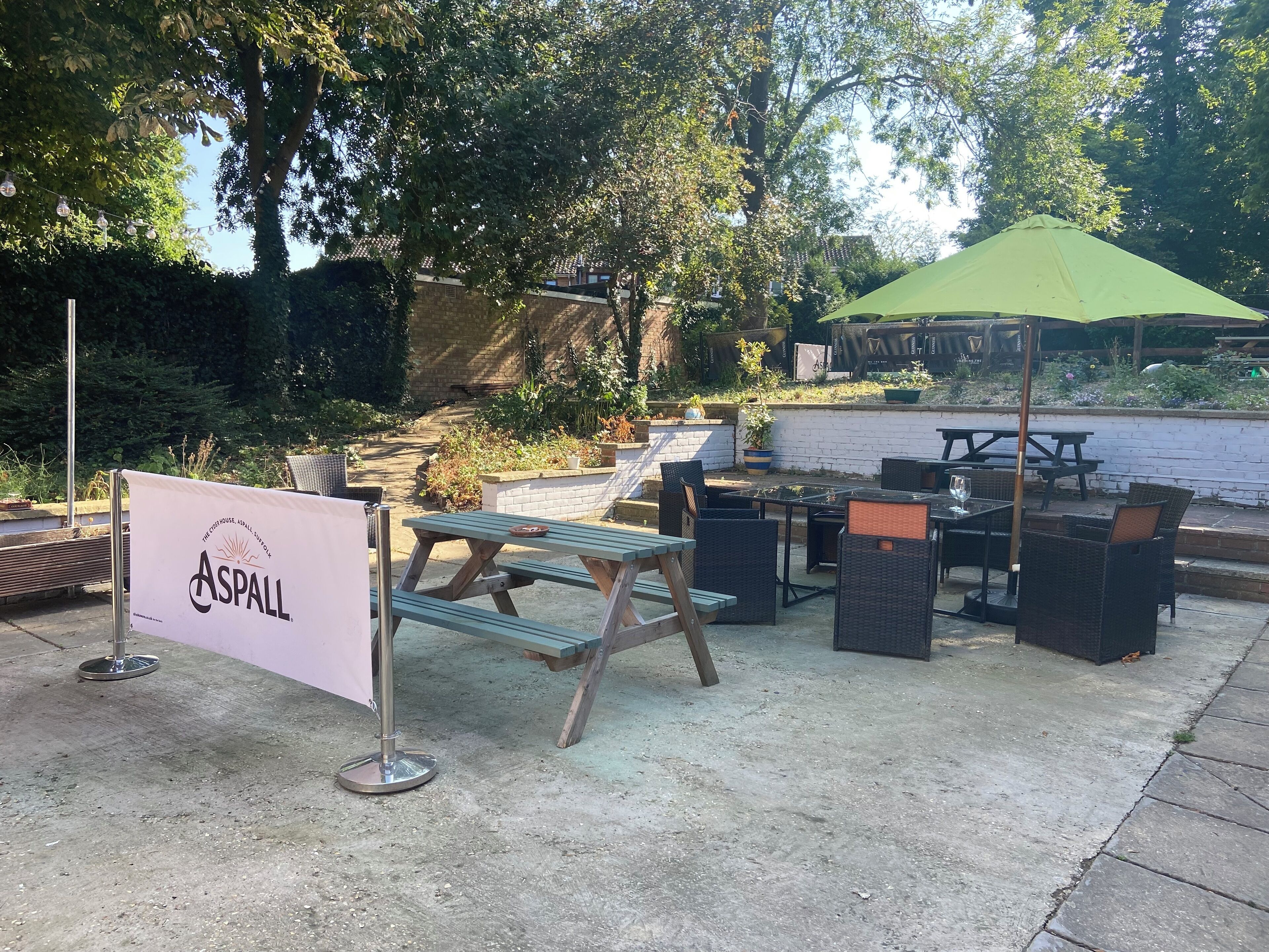 Een terras buiten met picknicktafels en stoelen, een groene parasol en een bordje met 'Aspall'.