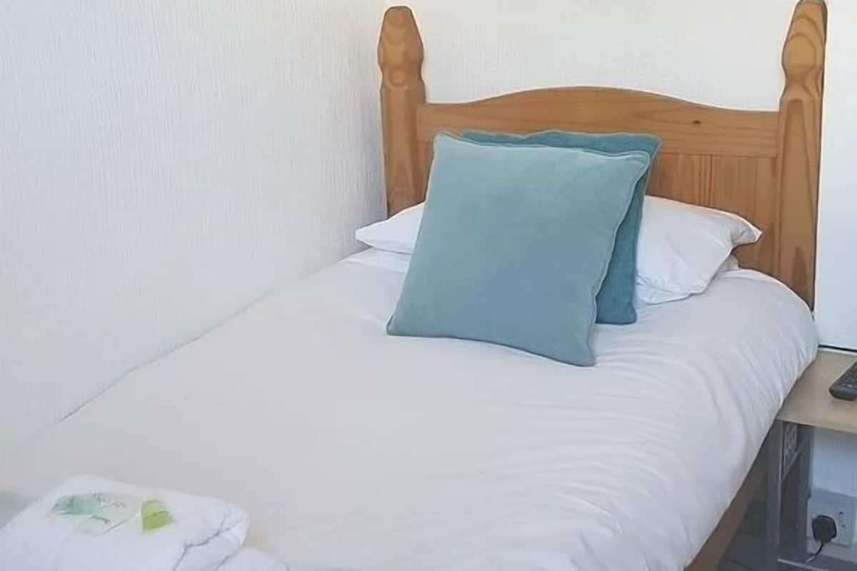 A single bed with a wooden headboard, white bedding, and a blue pillow. There is a folded white towel on the bed and a brown blanket at the foot.