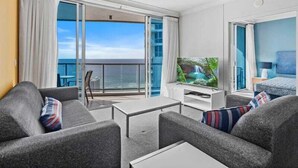 A modern hotel room with a balcony, a flat-screen TV, grey sofas, and a small table. - Ocean View Rental Surfers Paradise (Surfers Paradise)