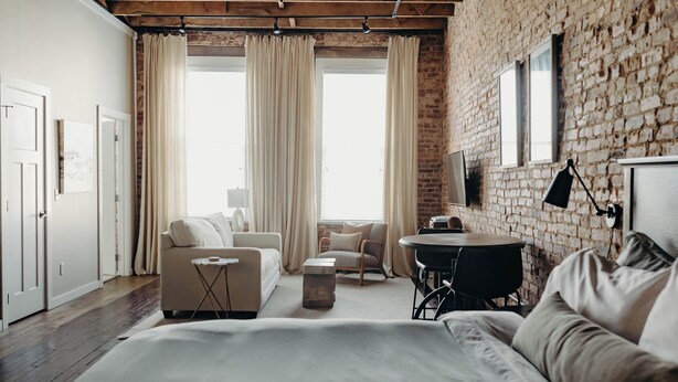 Een moderne slaapkamer met een groot bed, een sofa, een kleine tafel en een stoel. Er is een raam met gordijnen, een bakstenen muur en een flatscreen televisie die aan de muur is bevestigd.