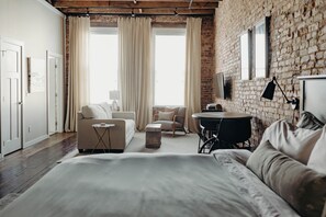 Un dormitorio moderno con una cama grande, un sofá, una mesita y una silla. Hay una ventana con cortinas, una pared de ladrillo y un televisor de pantalla plana montado en la pared.