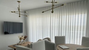 Un moderno salón con comedor, que cuenta con un amplio ventanal con cortinas translúcidas, un televisor de pantalla plana montado en la pared y una lámpara de araña original.