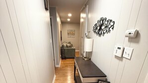 A narrow hallway with wooden flooring, a dark table with a lamp, a 'WELCOME' sign, and a wall-mounted thermostat.